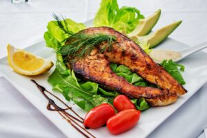 Delicious grilled salmon steak served with avocado, lettuce, cherry tomatoes, and lemon slice on a white plate.